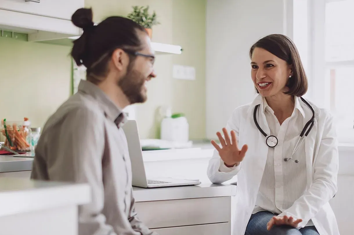 Physician, a white woman in a white coat, having a happy conversation with a patient, a Hispanic man with a top knot.