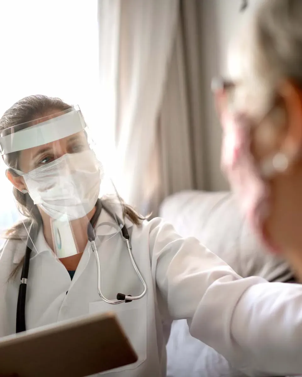 A physician, a white woman, in white coat, face mask and shield, talking to a patient, a senior Hispanic woman, in her home.