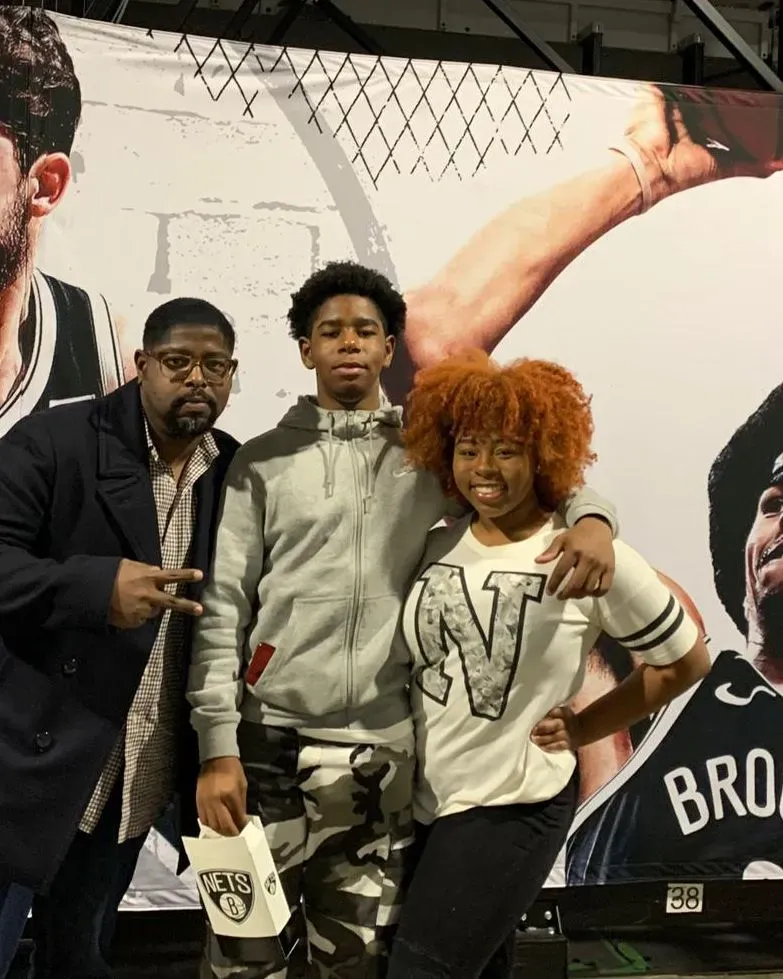 Neuro-Oncologist Dr. Phillips, a black man, hugging his two young adult children in front of a mural at the Barclays Center.