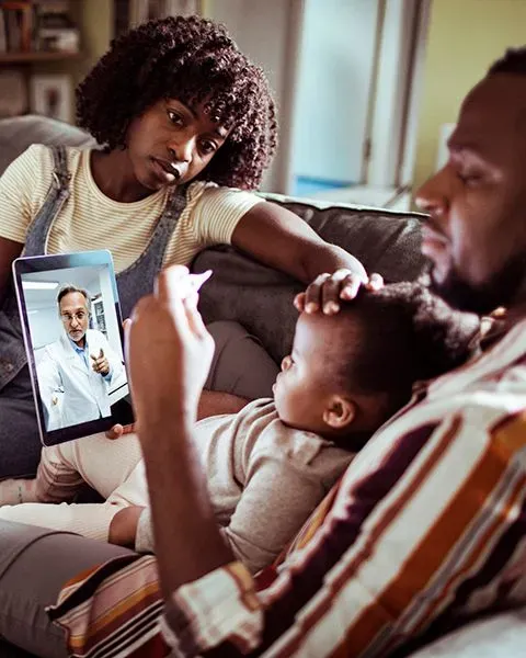 A Black family: a father, mother and baby on the couch talking to the pediatrician on a tablet.