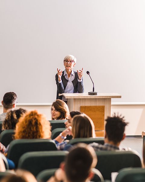 A Doctoral Faculty Member, a white woman with short white hair, teaching in front a class of students. 