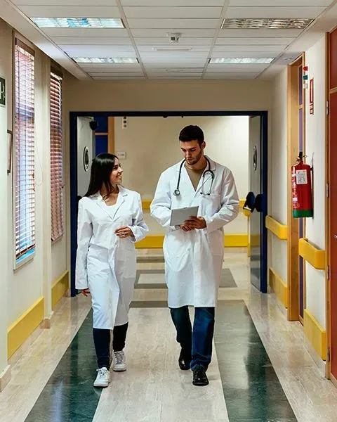 Two physician assistants walking down a hospital hallway, both in white coats, one a Hispanic woman, the other a Persian man.