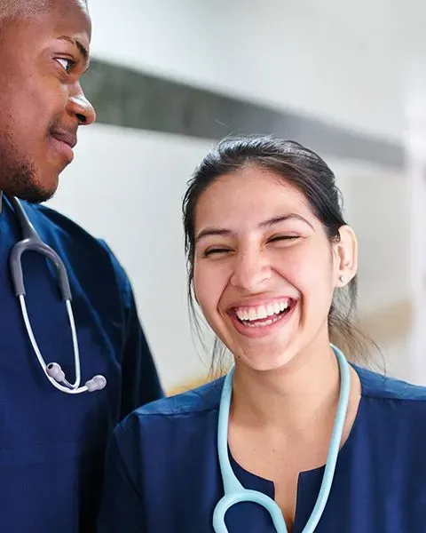 Two physicians in blue scrubs with stethoscopes laughing together, one is Black man, the other an Asian woman.