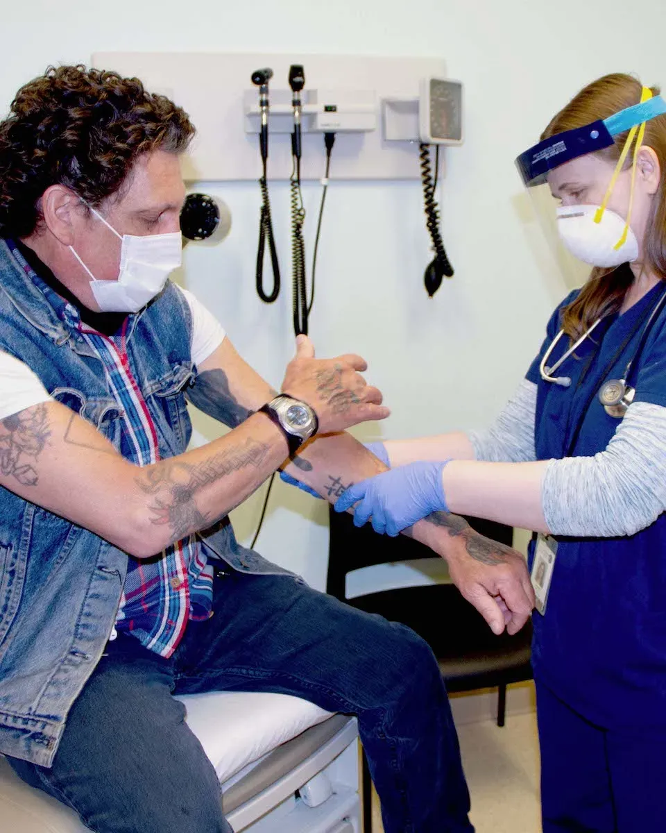 A physician, a white woman, in PPE examining the tattooed arm of a white masked-man in a Central City Concern clinic.