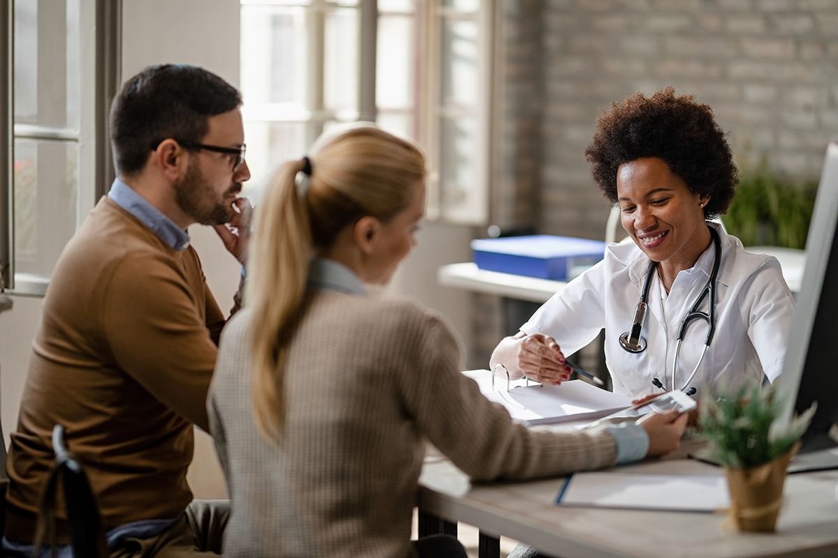 PS&D hired OB-GYN, a Black woman with a white coat, talking to a husband and wife.