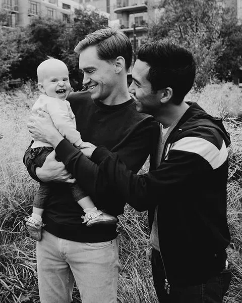Cardiologist Dr. Jason Bensch, a white man, smiling and holding his baby daughter along with Dr. Jason Heino, his husband.