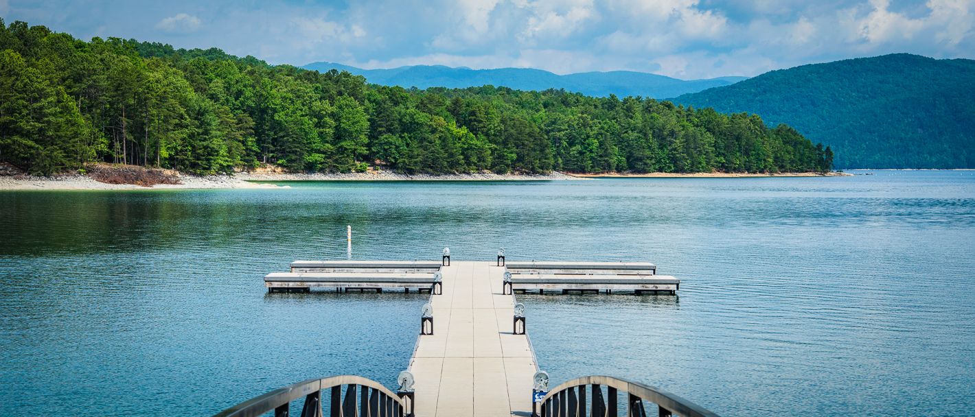 Sunny day at Lake Jocassee, SC. Dock stretches over blue water, framed by lush forest backdrop.