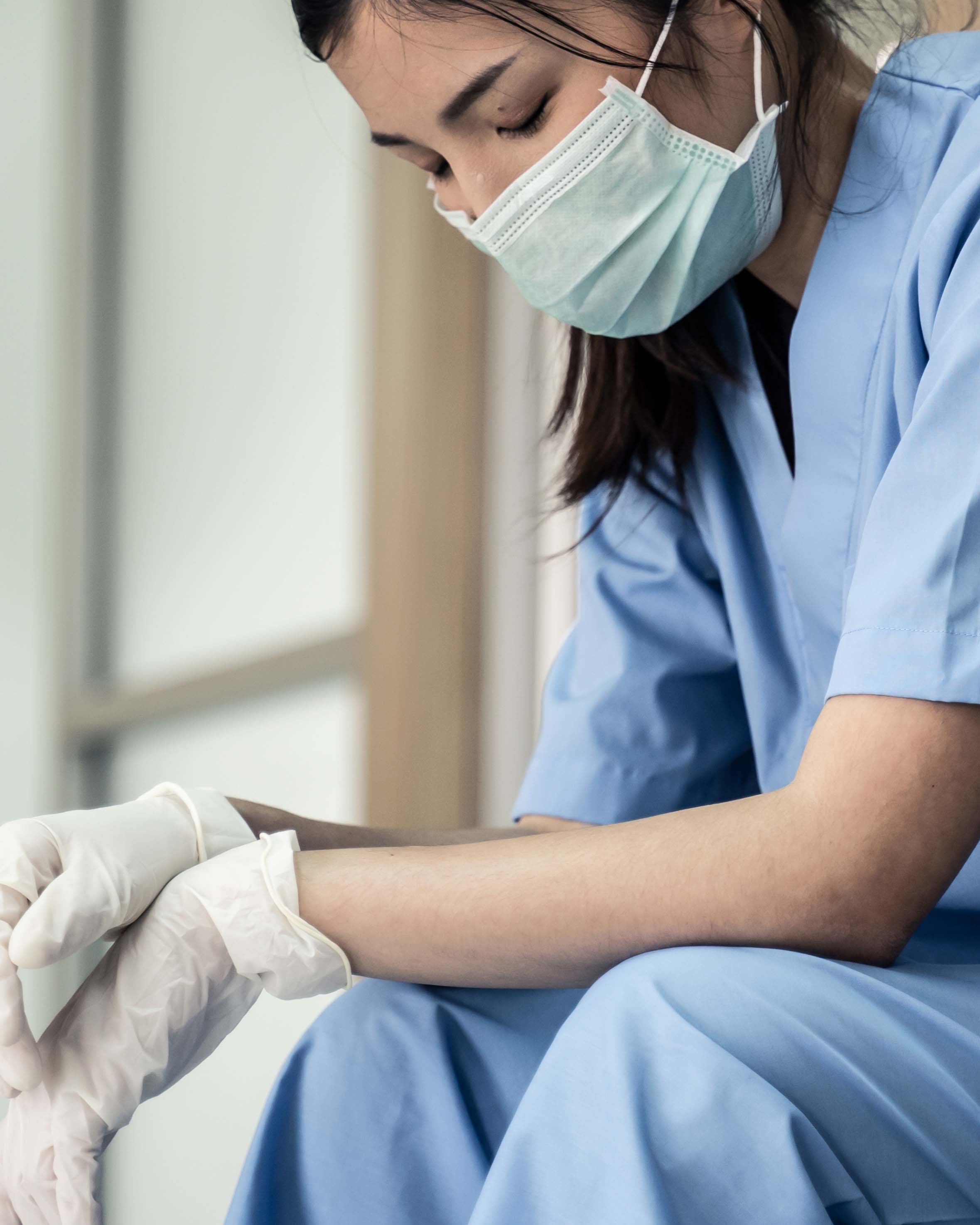 A physician, an Asian woman, in blue scrubs, a face mask and gloves, sitting down with her eyes closed, tired from her shift.