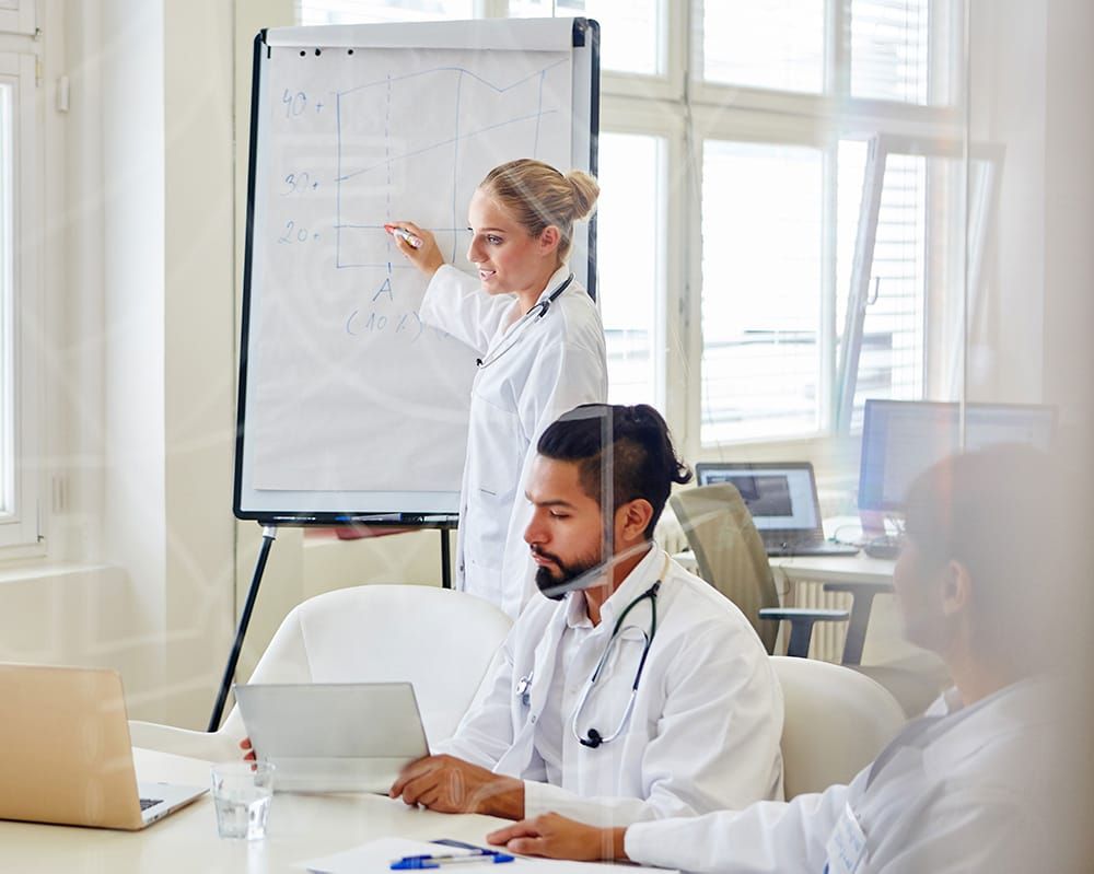 A physician, a white woman, presenting on an easel to her colleagues, a Hispanic man and an Asian woman, all in white coats.