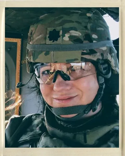 U.S. Army physician Dr. Wendy Miklos, a white woman, smiles wearing full army equipment with helmet, sitting in a helicopter.