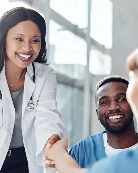 A physician, a Black woman, in a white coat, shaking hands of medical residents in scrubs, a Black man and white woman.