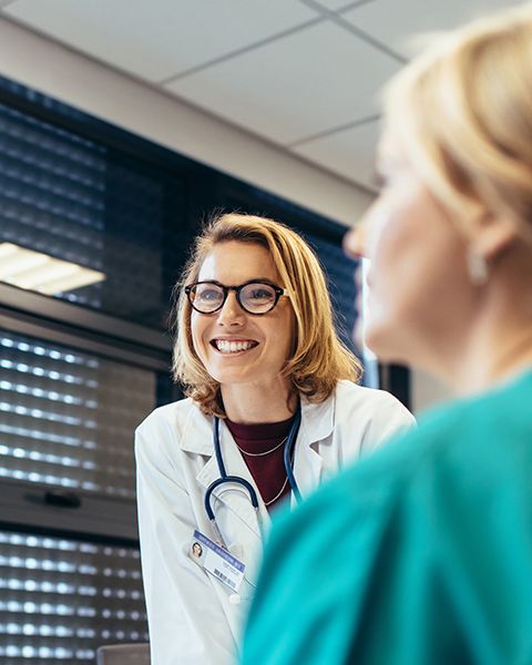 A physician, a white woman, wearing a white coat, glasses, smiling as she leads her team at a conference table.