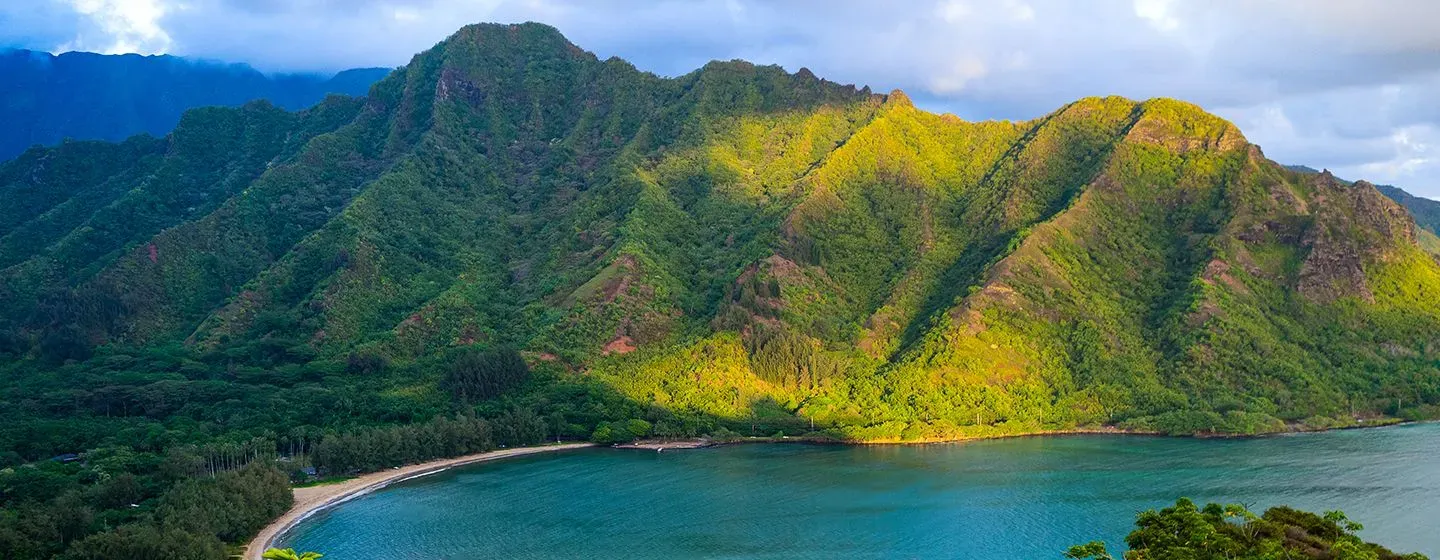 Lush, green mountains in front of the ocean with the sun peaking through the clouds in Honolulu, Hawai'i.