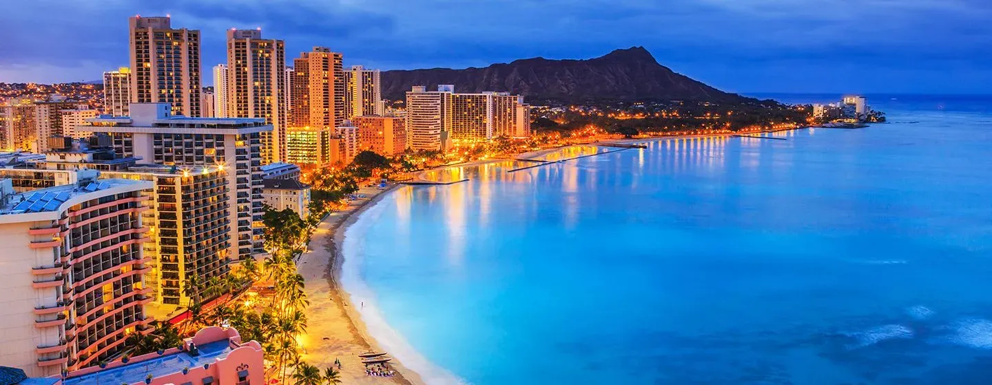 Night skyline view of downtown Honolulu, Hawai'i with glowing city lights in front of the ocean.