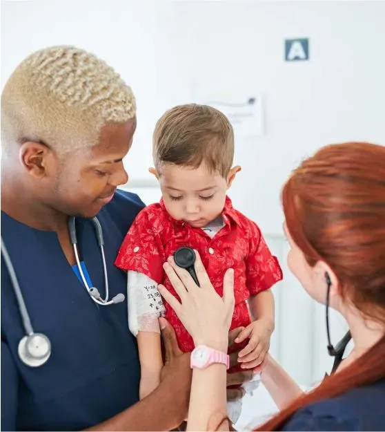 A physician, a Black man, holding a toddler while another physician, a white woman, uses a stethoscope. 