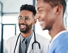 Two Family Medicine physicians, both young Black men, sharing a laugh, one in a white coat and the other in blue scrubs.
