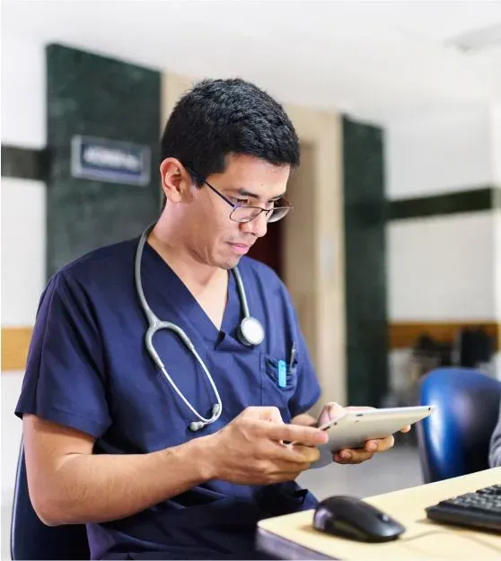 A physician, an Asian man, in blue scrubs and stethoscope looking at a tablet.