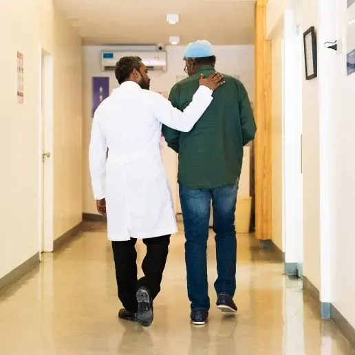A physician with his arm over his patient's shoulder as they both walk down a hallway together, both are Black.