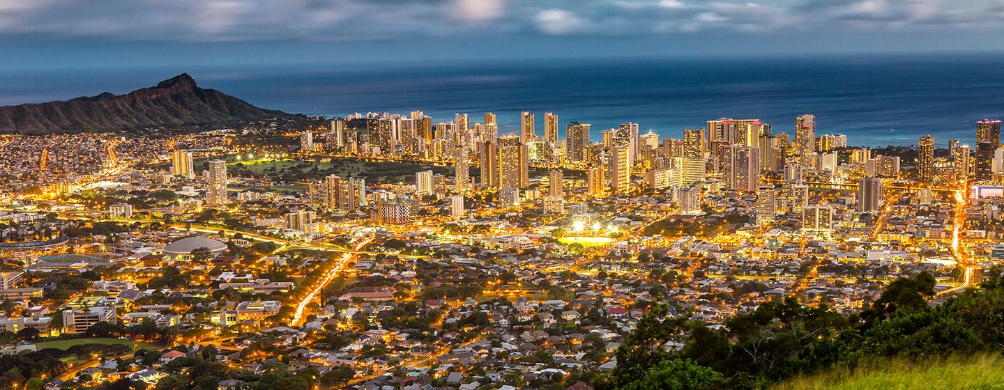 Aerial view of a downtown city in Hawai'i with glowing city lights with the ocean in the background.