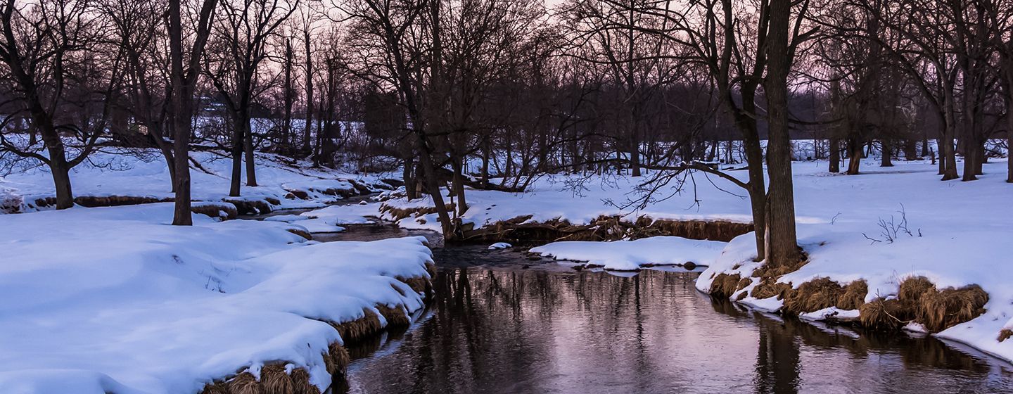 Snowy bank in front of a river surrounded by trees with no leaves in Pennsylvania.