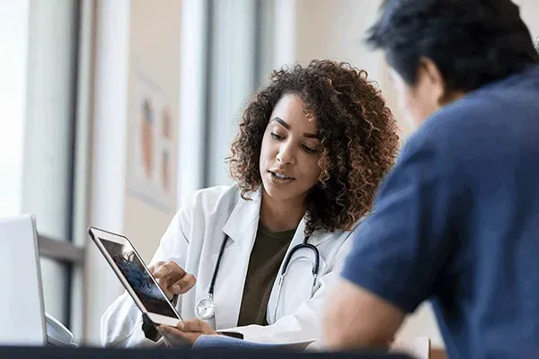 A Cardiologist, a Black woman in a white coat, using a tablet to discuss care plan with her patient, a Hispanic man.