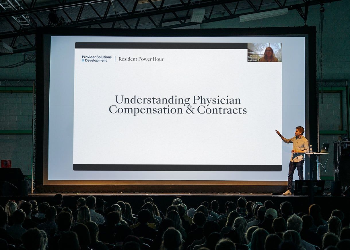 A white man presenting to a crowd in front of a big screen that says, "Understanding Physician Compensation and Contracts."