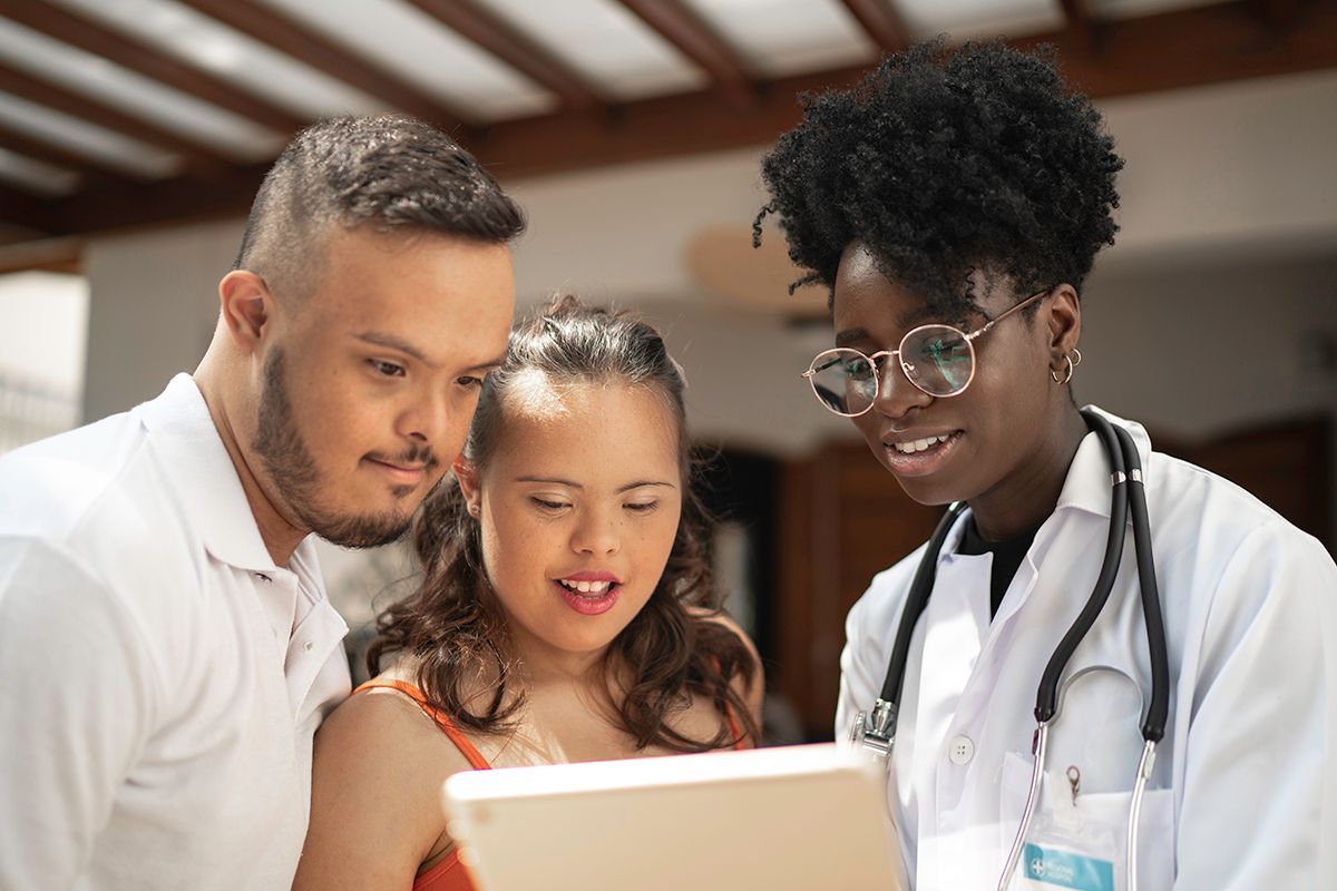 A physician, a Black woman in a white coat, shares medical file with patient, a young Southeast Asian man with down syndrome.