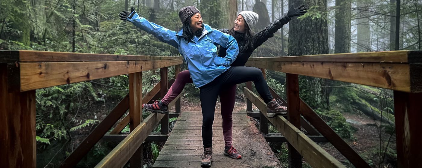Two physicians, both Asian women, in athleticwear on a hike, posing and smiling standing on a wooden bridge in the forest.