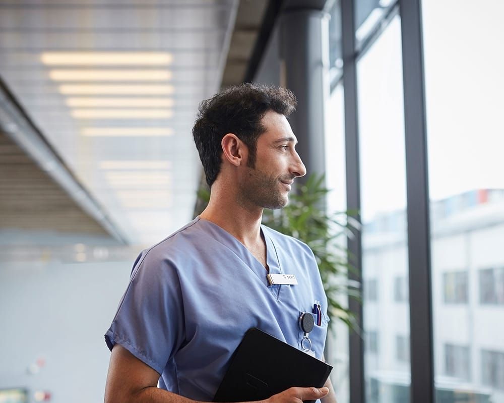 A physician, a Persian man, in blue scrubs, looking out the window contemplating his career.