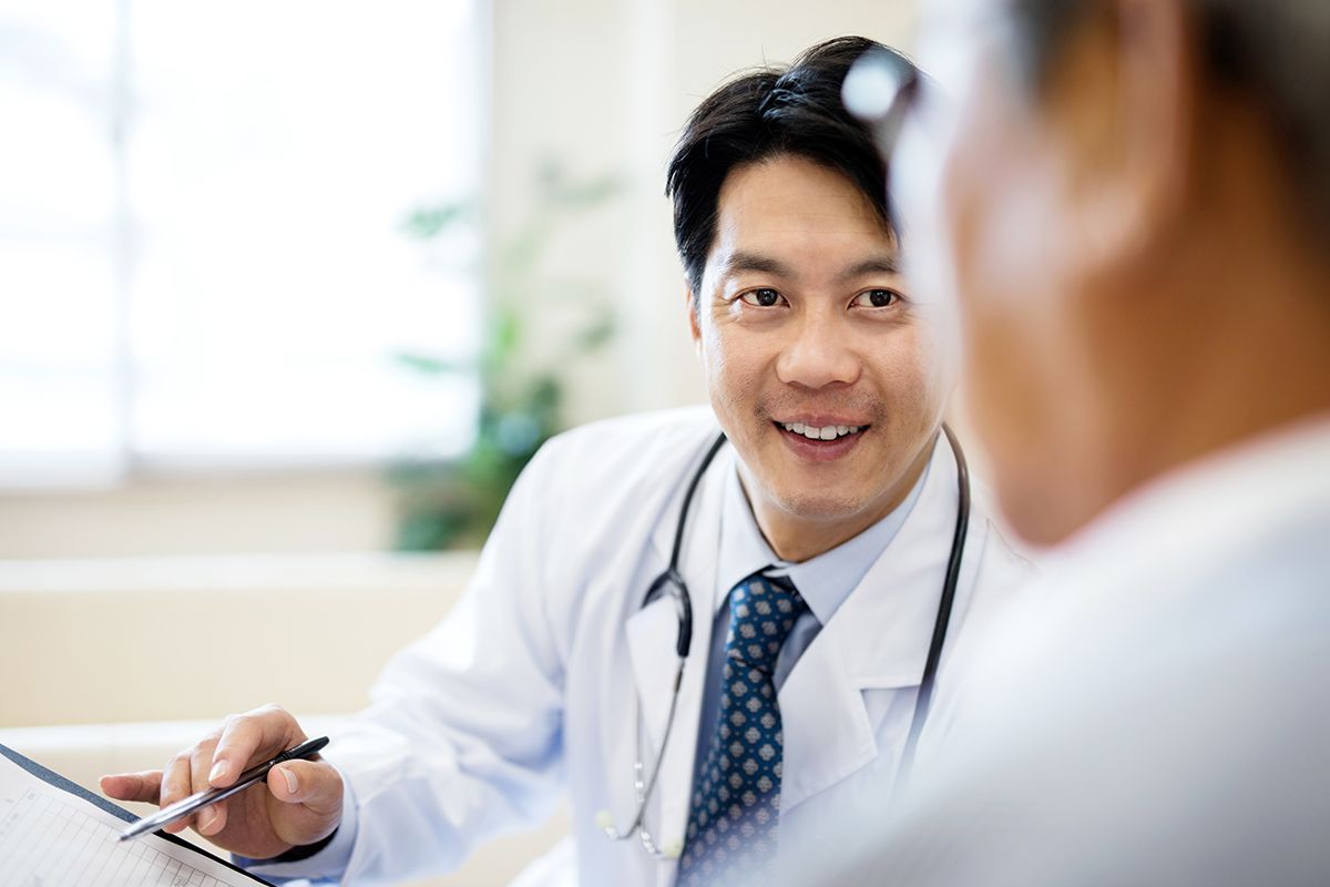 A Physician, an Asian man, in a white coat smiling and pointing to a file, while talking with a patient, an Asian older man.