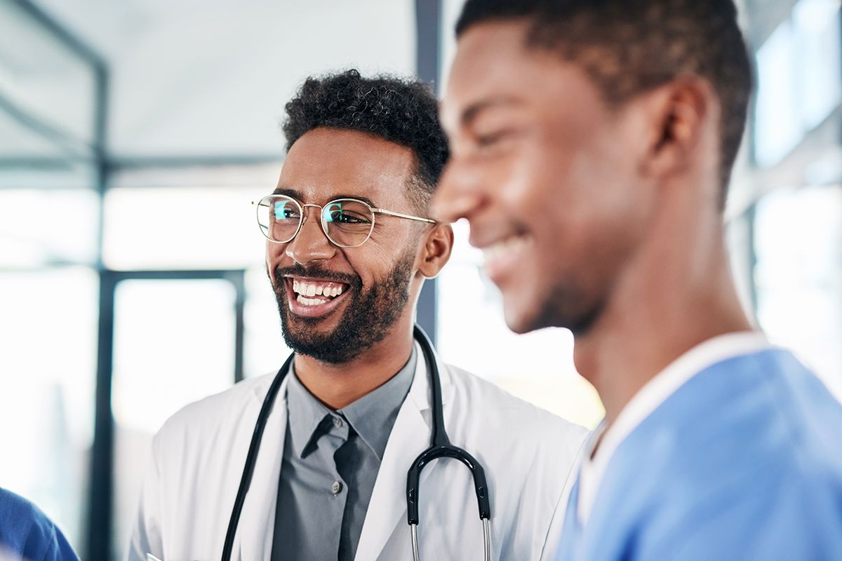 Two Family Medicine physicians, both young Black men, sharing a laugh, one in a white coat and the other in blue scrubs.