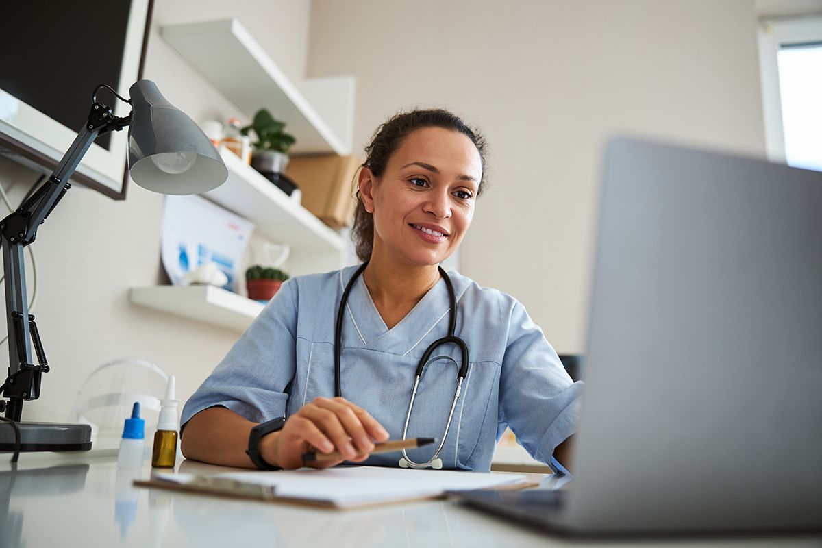 A Gastroenterologist, a young Black woman, in blue scrubs and stethoscope, at her laptop reviewing client files and smiling.