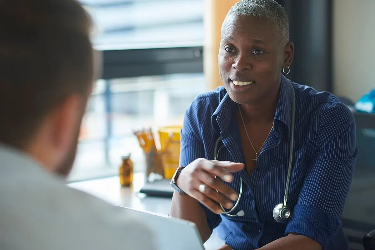 A Neurologist, a Black woman, in a blue oxford shirt and stethoscope, talking with her patient, a white man.