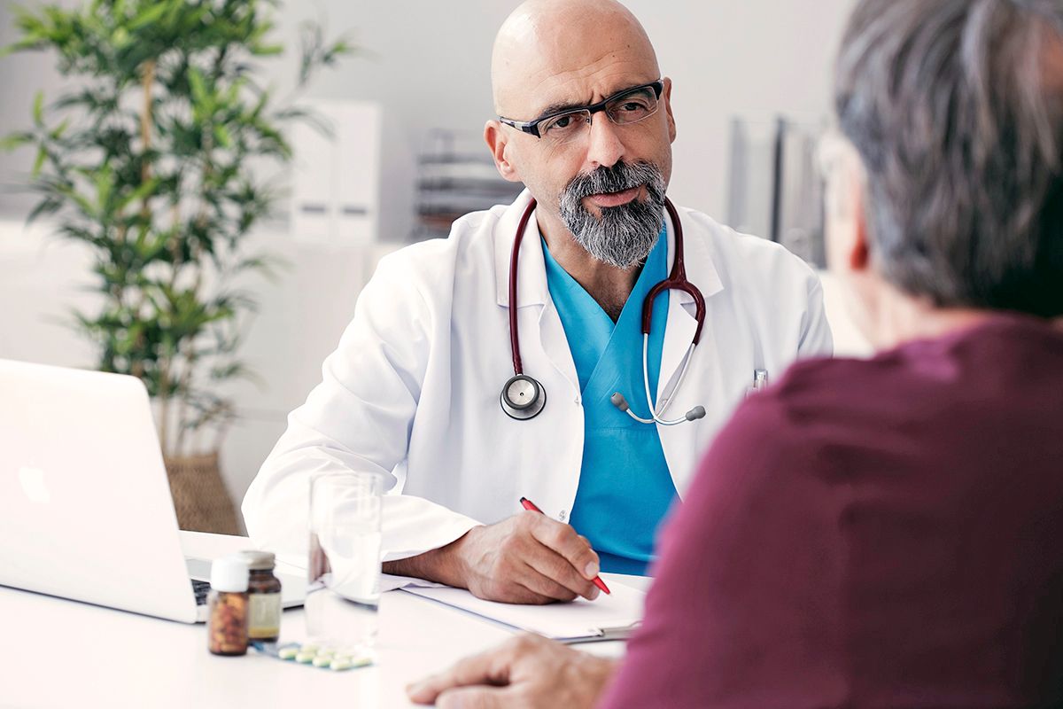 An Orthopedic Surgeon, a white man, in a white coat and stethoscope, sitting down talking with a patient, a senior Asian man.