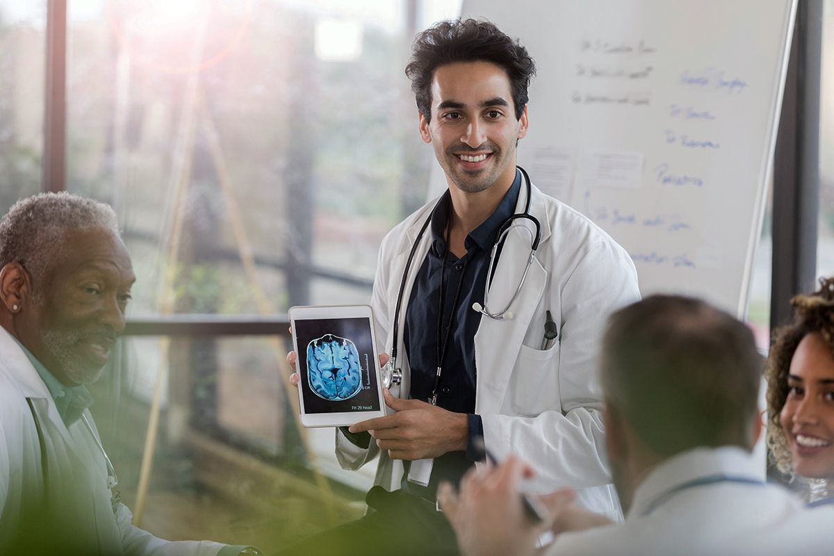 A Neurologist, an Indian man, in a white coat, consulting a group of multiethnic neurologists, a brain scan on a tablet.