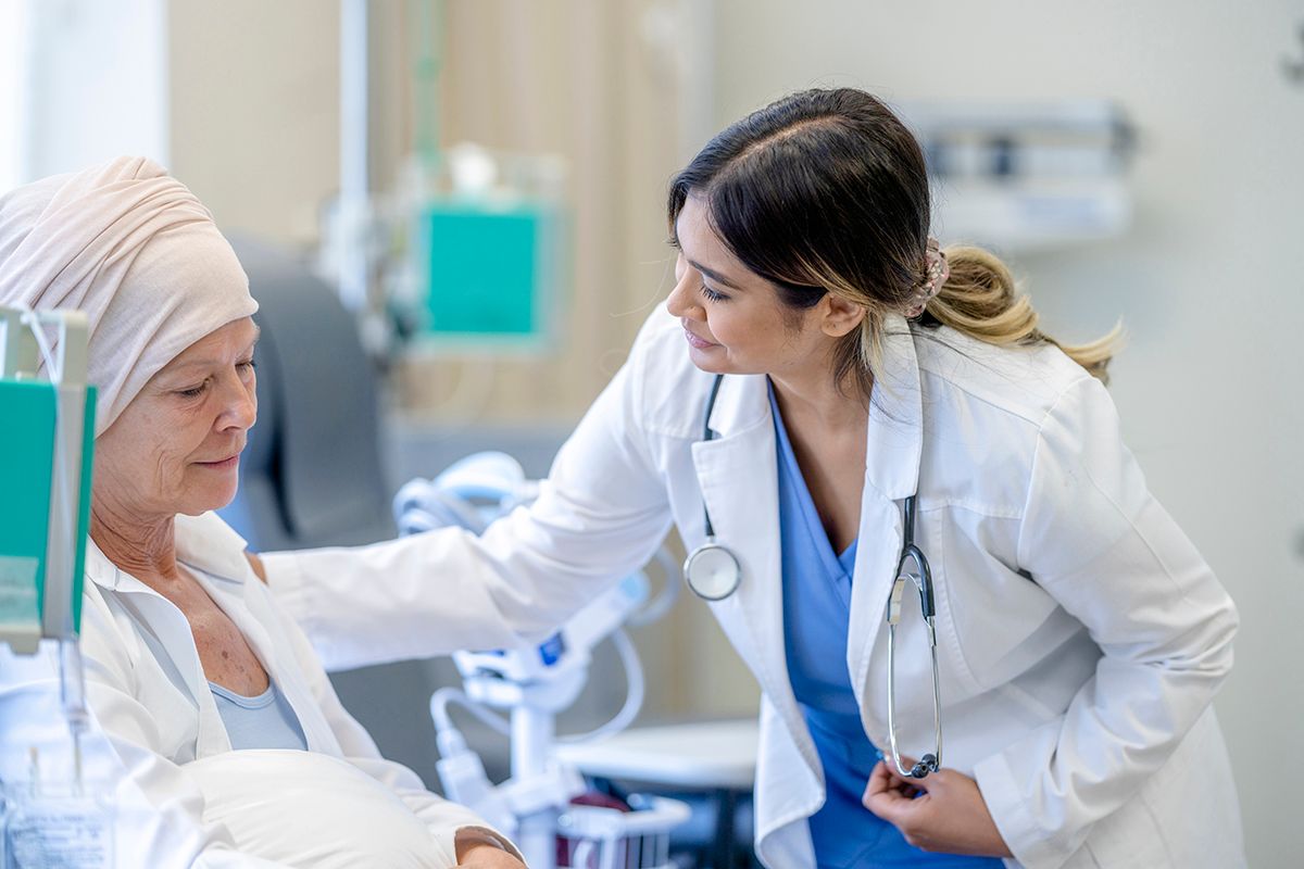 An Oncologist, a Hispanic woman, in a white coat, with her arm gently on her patient, a white woman, sitting a hospital bed.