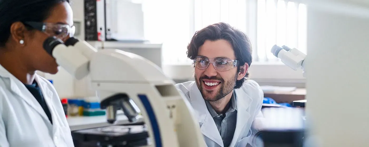 An Oncologist, an Indian woman, looking at a microscope, while a colleague, a white man, smiles and sits next to her.