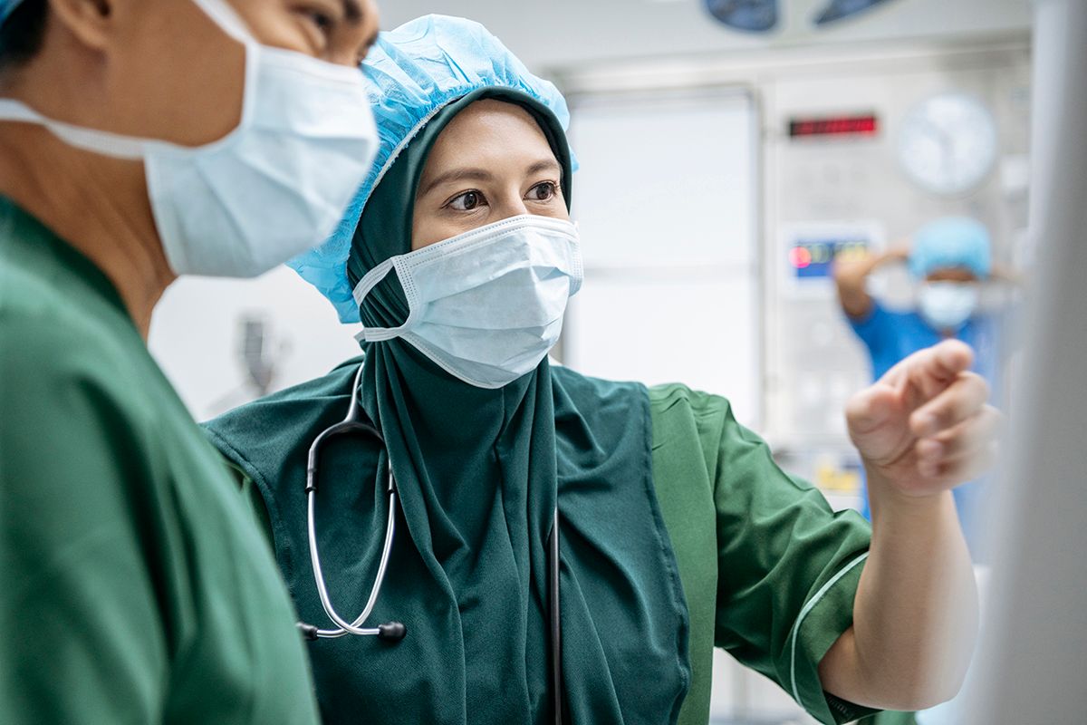 An Orthopedic Surgeon, an Asian woman wearing a hijab, reviewing a monitor with a colleague, a Hispanic man, both in PPE.