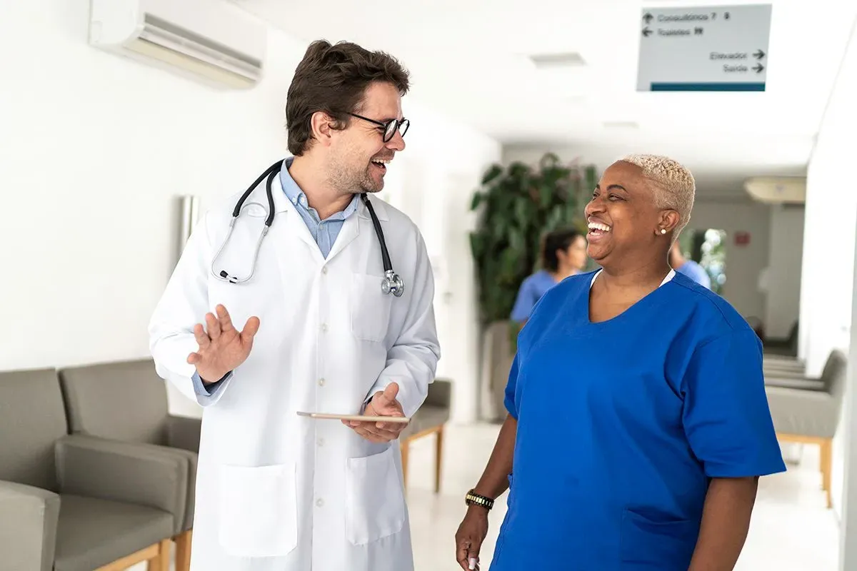 An Orthopedic Surgeon, a Black woman, in blue scrubs shares a laugh with a fellow surgeon, a white man in a white coat.