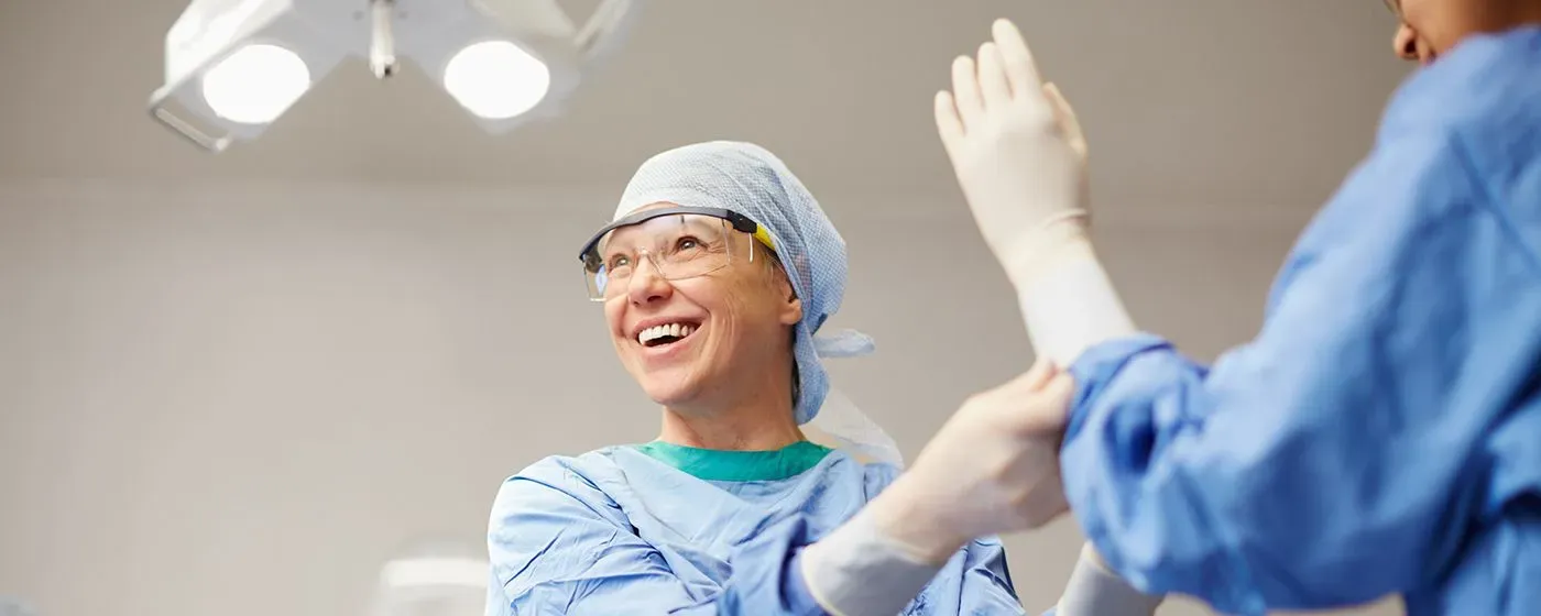 An Orthopedic Surgeon, a white woman, smiles, preps for surgery, helps a colleague, an Indian woman, put on surgical gloves.