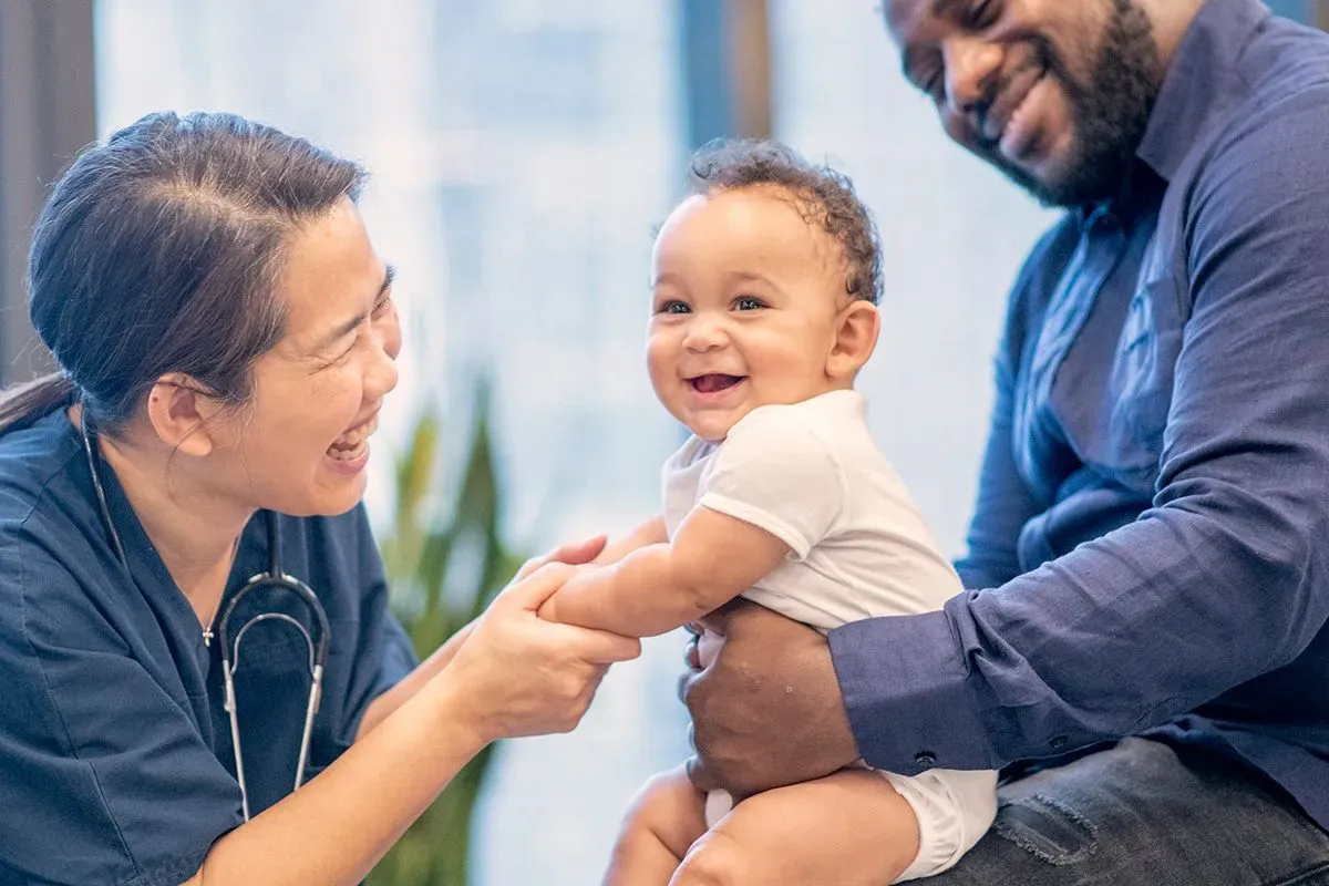 A Pediatric physician, an Asian woman, making her patient laugh, a Black baby girl, sitting on top of her dad's lap.