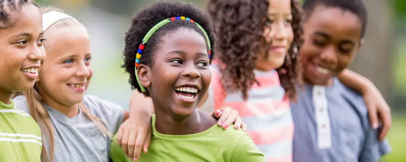 A multiethnic group of children, a young Black girl at the center in natural curls smiling and hugging a white young girl.