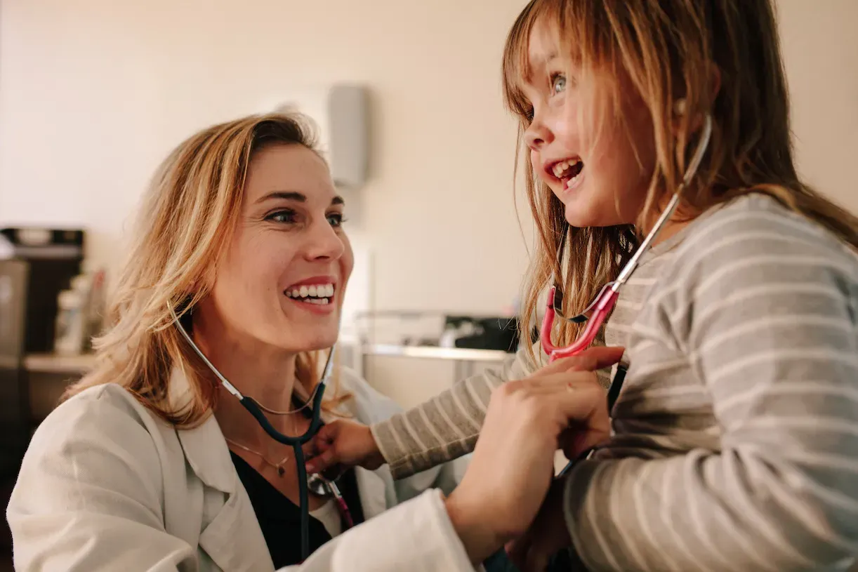 Female doctor checks the heart of a young girl with a stethoscope while both are smiling.