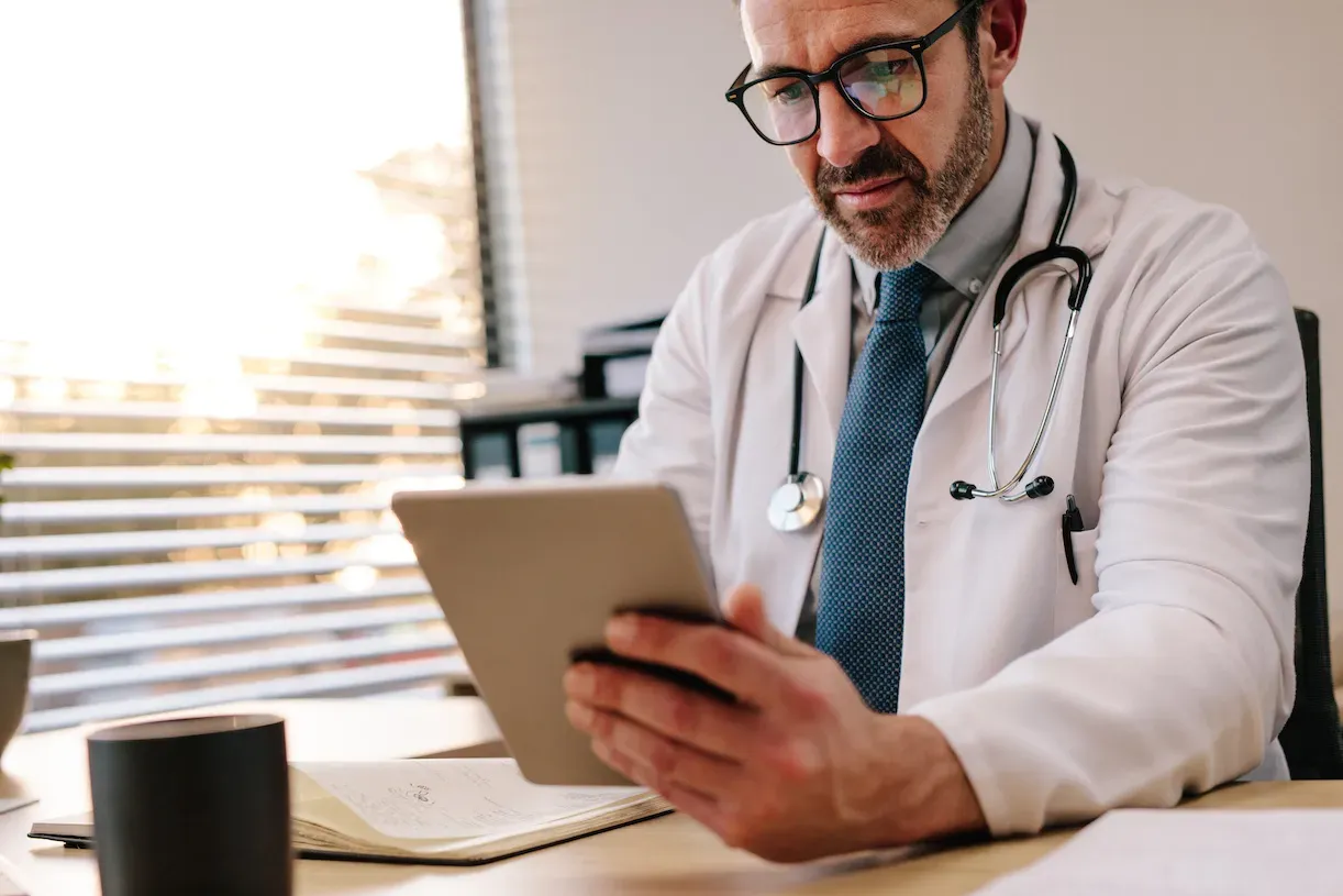 A male doctor is looking intently at a tablet he is holding while sitting at his desk.