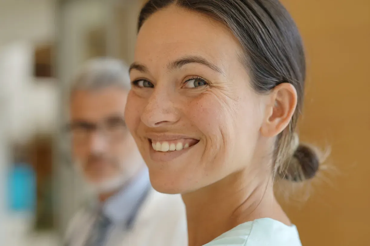 Female looking directly at the camera and smiling. There is a male in the background also looking at the camera.