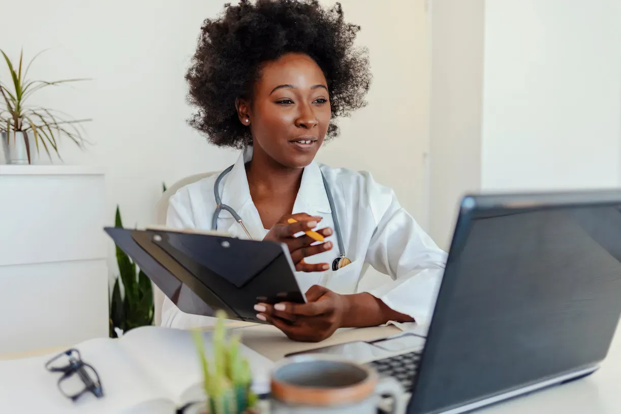 A female doctor is holding a clipboard while looking at her laptop at her desk.