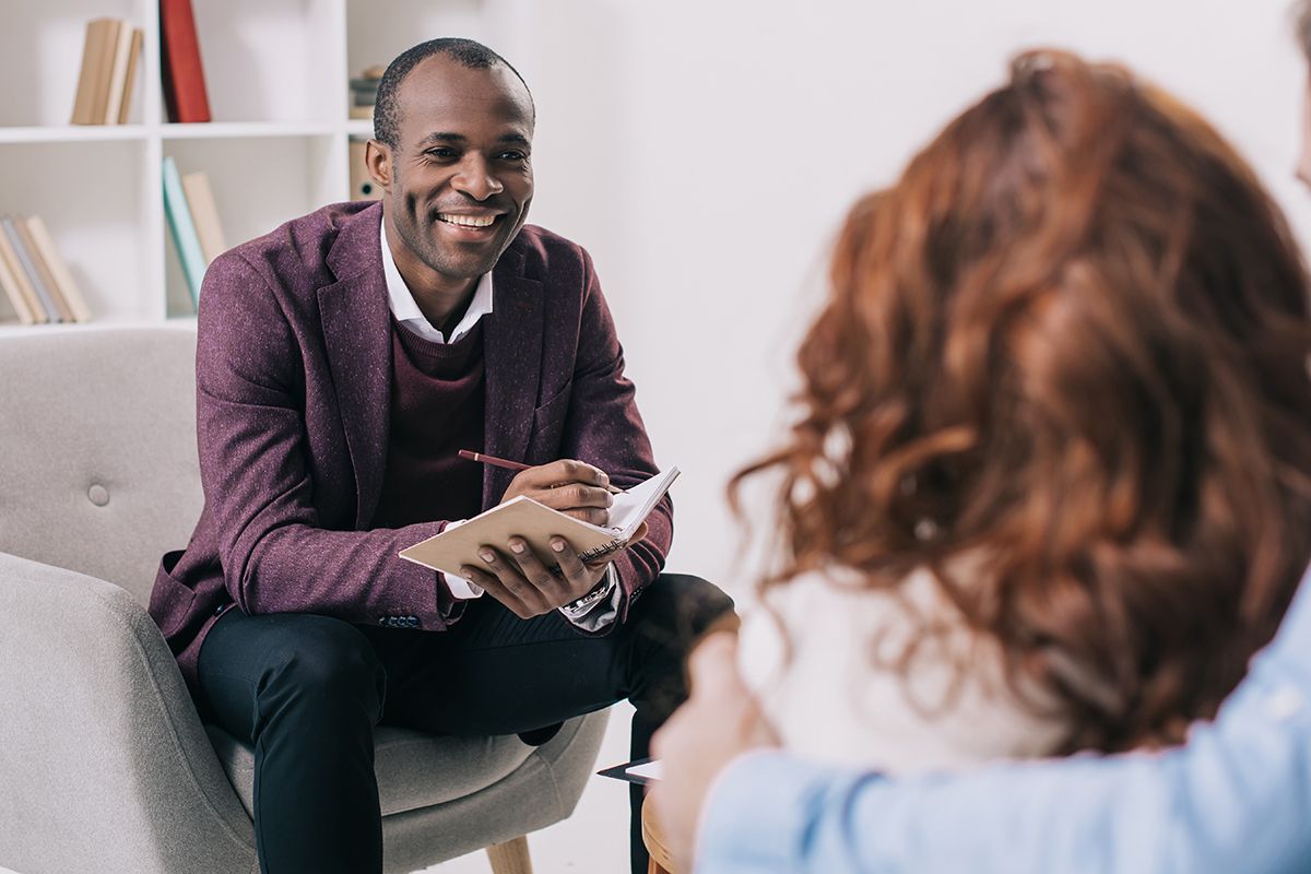 Psychiatrist, a Black man sitting in an armchair, takes notes and smiles at his patient, a curly red-haired girl with her dad’s arm around her.