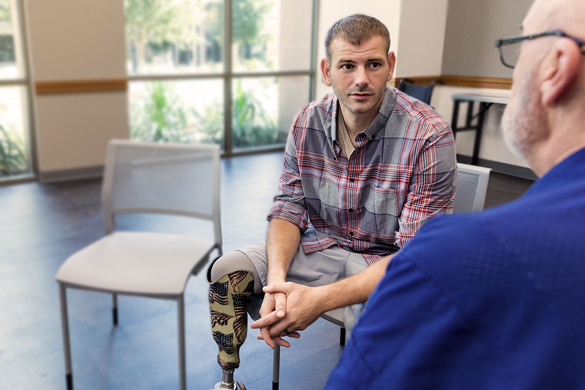 A veteran, a white man in a plaid top and khaki shorts, with prosthetic leg, talking with a Psychiatrist, a white senior man.