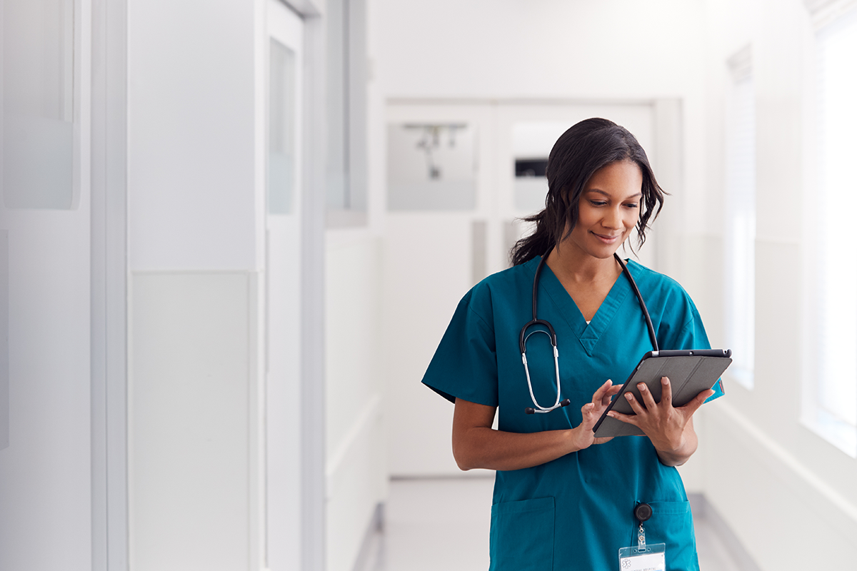 A physician, a Black woman, in blue scrubs, smiling, while holding a medical chart, walking down a hospital hall.